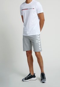 Man med vit Hummel-t-shirt, grå Hummel-short med chevronmönster och svarta och grå träningsskor som står mot en enkel bakgrund.