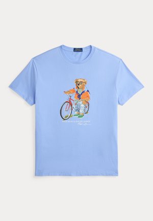 Camiseta azul clara con un gráfico de un oso que lleva gafas de sol, una chaqueta naranja, vaqueros y zapatos verdes, de pie junto a una bicicleta roja.