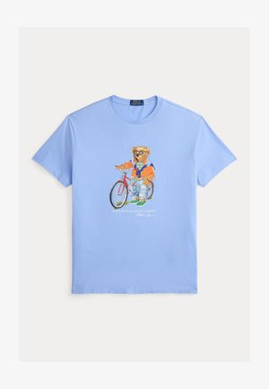 Camiseta azul clara con un gráfico de un oso que lleva gafas de sol, una chaqueta naranja, vaqueros y zapatos verdes, de pie junto a una bicicleta roja.