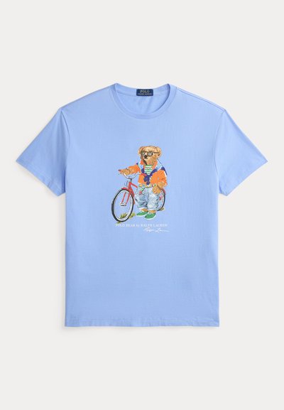 Ανοιχτό μπλε μπλουζάκι με γραφικό ενός αρκούδου που φοράει γυαλιά ηλίου, πορτοκαλί μπουφάν, τζιν και πράσινα παπούτσια, που στέκεται δίπλα σε ένα κόκκινο ποδήλατο.