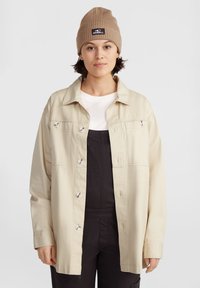 Chaqueta beige de botones con cuello y herrajes plateados, llevada sobre una camisa blanca y un overol oscuro, combinada con un gorro de punto acanalado en tono beige.