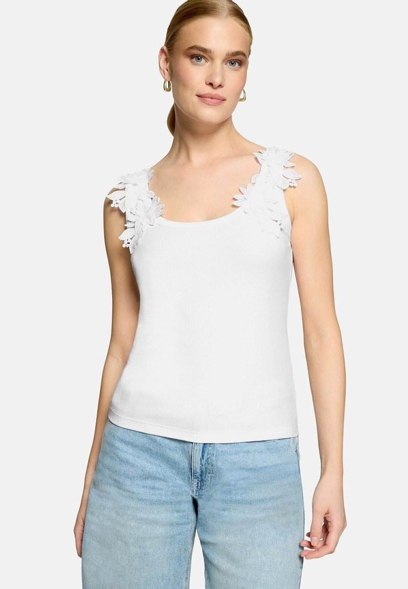 Femme portant un haut blanc sans manches avec des détails en dentelle florale sur les bretelles et un jean bleu clair, debout devant un fond uni.