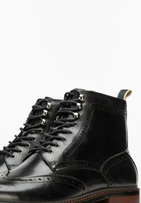 Botas de cuero negro con detalles de brogue, diseño de cordones, ojales de metal y acabado texturizado. Presentan un tacón apilado y costuras en contraste.