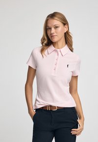 Polo de color rosa claro con cuello, que presenta una tapeta abotonada y un pequeño logotipo bordado en el lado izquierdo. Combinado con pantalones oscuros y un cinturón marrón.