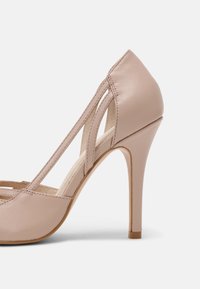 Chaussure à talons hauts en cuir beige avec un design à bride en T et talon fermé, présentant un talon aiguilles fin et une finition lisse.