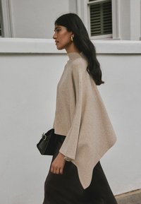 Top de punto beige texturizado con cuello alto y mangas anchas, combinado con una falda oscura y un pequeño bolso negro.