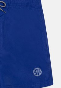 Pantaloni da bagno blu realizzati in materiale leggero. Vita elasticizzata con cordino, tasca laterale e un logo bianco di una palma vicino all'orlo.