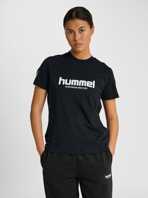 Ung kvinde med mørkt hår samlet bagpå iført en sort Hummel T-shirt og sorte bukser, stående med hænderne i lommerne mod en ensfarvet baggrund.
