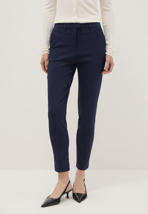 Woman wearing fitted navy ankle-length pants, white ribbed buttoned top, and black pointed-toe slingback heels standing against a plain background.