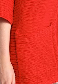 Czerwony sweter z dzianiny z poziomymi żebrami, krótkimi rękawami oraz dużą kieszenią naszywaną z przodu. Teksturowany materiał dodaje głębi.