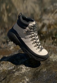Bota de caminhada bege com um colarinho alto, detalhes cinza e ilhós de metal. Apresenta uma sola acolchoada e uma superfície texturizada para melhor aderência.