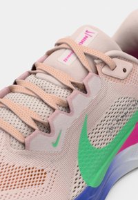 Nike kurpes ar bēšu sieta augšdaļu, rozā un zaļiem akcentiem, teksturētām šņorēm un daudzkrāsainu zoli ar ziliem, zaļiem un rozā sekcijām.