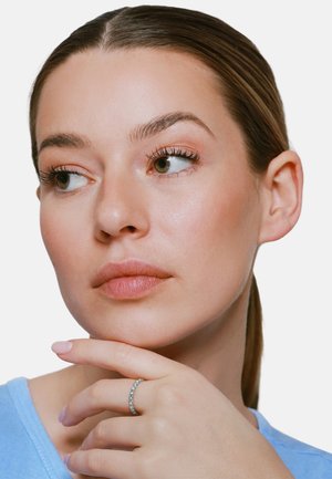 Donna dalla pelle chiara e capelli castani guarda di lato, appoggiando il mento su una mano con un anello d'argento tempestato di diamanti, indossa un top azzurro chiaro.