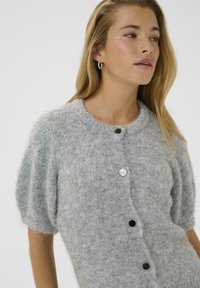 Cardigan en tricot gris avec des manches courtes bouffantes, présentant un col rond et une fermeture à boutons sur le devant. Tissu texturé avec des boutons argentés et noirs.