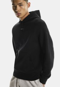 Zwarte hoodie gemaakt van zachte stof, met een voorzak, een capuchon met trekkoord en een minimalistisch ontwerp. Geaccentueerd met een klein merklabel.