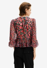 Blusa floreale trasparente con fiori rossi su sfondo nero, maniche a sbuffo e orlo sfrangiato. Abbinata a pantaloni cargo neri.