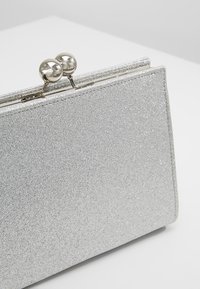 Pochette rectangulaire argentée pailletée avec fermeture à boule métallique sur une surface blanche.