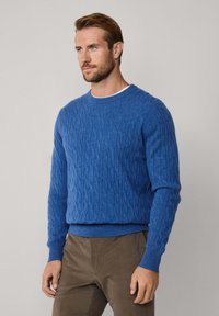 Suéter azul de punto trenzado con cuello redondo, patrón texturizado y puños y dobladillo acanalados. Combinado con pantalones marrones.