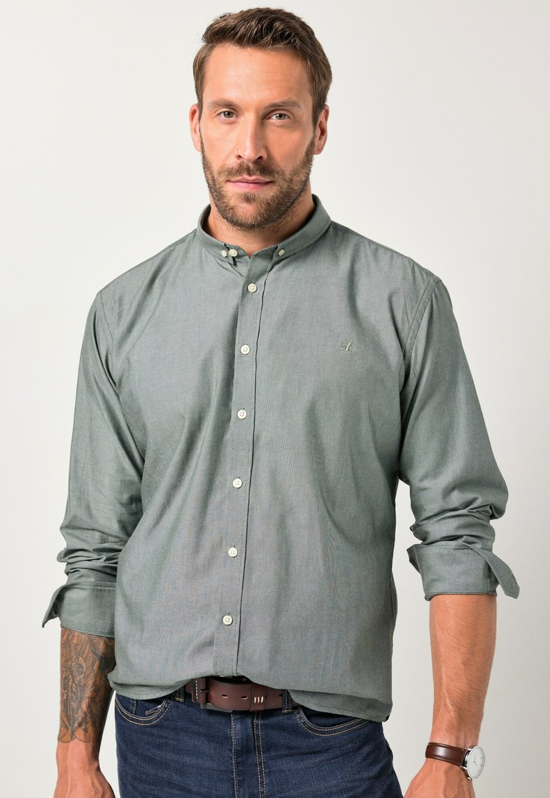 Uomo con la barba che indossa una camicia verde con i polsini arrotolati, jeans scuri, una cintura marrone e un orologio da polso, in piedi contro uno sfondo semplice.