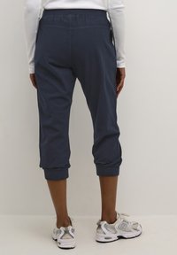 Pantalon court en coton bleu marine avec taille élastique et ourlets retroussés, associé à un haut blanc à manches longues et des chaussures de course grises.