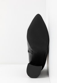 Semelle d'une botte noire à bout pointu avec talon en caoutchouc texturé et avant-pied, positionnée debout contre une surface blanche à angles.