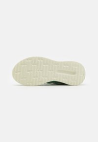 Semelle de chaussure en beige clair avec surface texturée et rainures. Tige en tissu vert partiellement visible, présentant un design rembourré et de soutien.