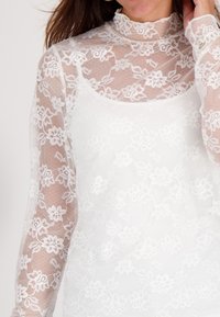 Witte kanten top met lange, doorzichtige mouwen met bloemmotief en een hoge kraag. Bevat een gladde, effen onderlaag voor bedekking.