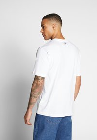 Biała bawełniana koszulka z krótkimi rękawami, z małym czarno-zielonym logo na karku. Noszone są do niej dżinsowe szorty.