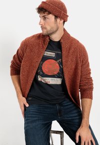 Rostfarbener Strickcardigan mit einem gerippten Kragen, über einem schwarzen Grafik-T-Shirt mit Bergmotiv getragen, kombiniert mit dunkelblauen Jeans.