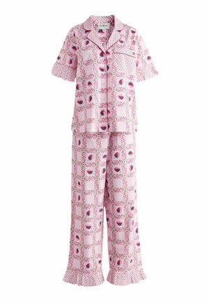 Nainen vaaleanpunaisissa kuviollisissa pyjamissa, joissa on röyhelöt hihoissa ja housujen lahkeissa, seisoo vuoteen vieressä, jonka pinkissä röyhelöisessä vuodevaatteessa ja pastellivärisissä verhoissa.