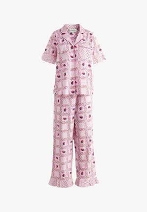 Nainen vaaleanpunaisissa kuviollisissa pyjamissa, joissa on röyhelöt hihoissa ja housujen lahkeissa, seisoo vuoteen vieressä, jonka pinkissä röyhelöisessä vuodevaatteessa ja pastellivärisissä verhoissa.