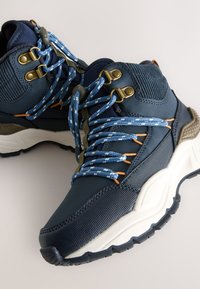 Marineblå vandrestøvler med struktureret stof, solide gummisåler, metaløjer og mønstrede snørebånd; forstærket hæl- og tådesign.