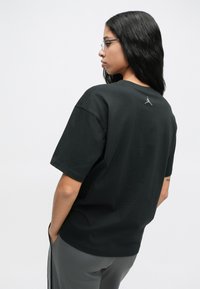 Γυναίκα με μακριά μαύρα μαλλιά, φορώντας γυαλιά και μαύρη φαρδιά μπλούζα με μικρό λογότυπο στο επάνω μέρος της πλάτης, με τα χέρια στις τσέπες, στραμμένη πλαγίως.