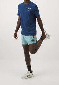 Camisa de manga corta azul con logo blanco, combinada con pantalones cortos azul claro que presentan un diseño negro y beige, y zapatos deportivos blancos con detalles.
