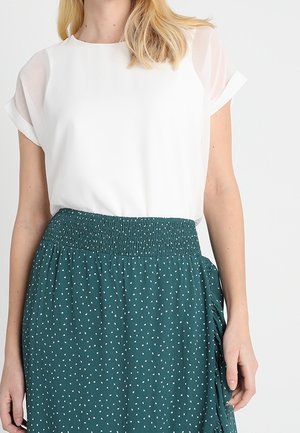 Kvinde iført en hvid kortærmet bluse, stukket ned i et højtaljet teal nederdel med hvide prikker og en rynket detalje i siden.