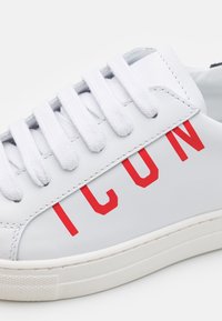 Baskets en cuir blanc avec surface texturée, lettrage rouge "ICON", six œillets blancs et semelle en caoutchouc blanche.