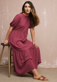 Staubrose Maxikleid mit Puffärmeln, gestuftem Rock und weichem Stoff; kombiniert mit braunen Riemchensandalen. Neutrale Kulisse verstärkt die Farben.