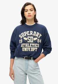 Sweatshirt en coton bleu marine avec un col rond, et le texte brodé en blanc et or "SUPERDRY 1984 ATHLETICS UNIV.DPT" ainsi que des poignets côtelés.