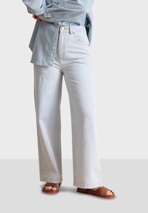 Jeans a gamba larga di un blu chiaro, realizzati in denim, con tasche frontali, accessori dorati e vita alta, abbinati a sandali marroni con cinturini.