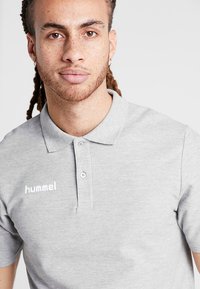 Grå polo-shirt lavet af bomuld med en to-knappet placket og en blød tekstur; har et hvidt "hummel" logo på venstre brystområde.