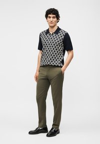 Νέος άνδρας που φοράει μαύρο-λευκό μπλουζάκι πόλο με σχέδιο, ελαιπράσινο παντελόνι και μαύρα επίσημα παπούτσια, στέκεται με το ένα χέρι στην τσέπη.