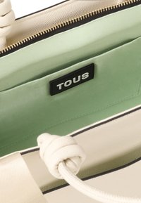 Sac à main beige avec une surface texturée et un intérieur en satin vert menthe. Il présente une étiquette noire avec "TOUS" et des poignées en corde blanche.