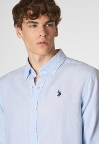 Un giovane con capelli disordinati indossa una camicia azzurra con bottoni che presenta un logo di un cavallo nero sul lato sinistro del petto.