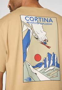 Μπεζ βαμβακερό μπλουζάκι με πολύχρωμο γραφικό ενός σκιέρ, βουνά και έναν κόκκινο ήλιο, με τα γράμματα "CORTINA" και "NEVER STOP EXPLORING".