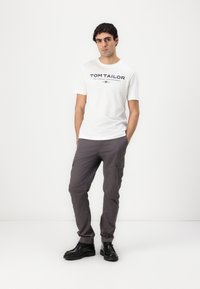 Hvid kortærmet t-shirt med "TOM TAILOR" trykt i sort, parret med grå cargo-bukser og sorte sko, som har flere lommer.