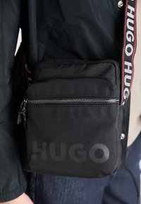 Borsa a tracolla nera in tessuto con chiusura con zip, caratterizzata da un audace logo "HUGO" e una tracolla regolabile con branding ripetuto.
