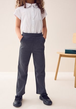 Kind steht mit Händen in den Taschen, trägt ein weißes kurzärmeliges Hemd, dunkelgraue Hose, schwarze Schuhe, neben einem Holzstuhl mit Büchern.