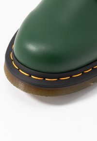 Gros plan sur l'avant d'une botte en cuir vert avec une semelle noire, des coutures jaunes et une semelle extérieure translucide marron sur fond blanc.