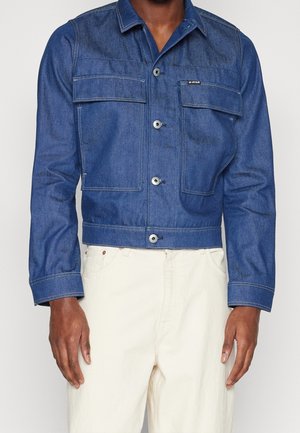 Man draagt een blauwe spijkerjasje met knoopsluitingen en klepzakken op de borst, gecombineerd met gebroken witte broek.