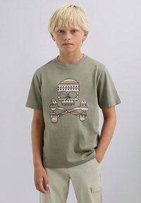 Camiseta de algodón verde oliva con un gran gráfico de calavera y huesos cruzados, con patrones geométricos en marrón, blanco y rojo. Mangas cortas.